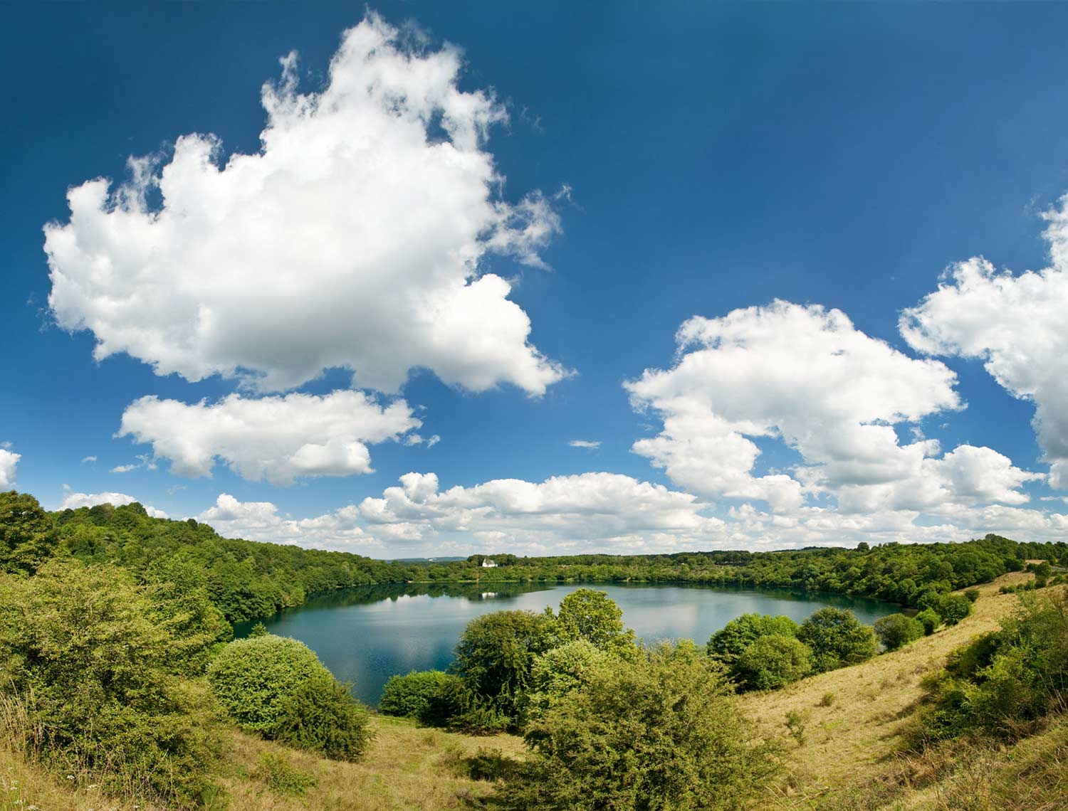 See, eingebettet in eine grüne Wiesen- und Buschlandschaft. Darüber ziehen große, weiße Wolken über den blauen Himmel