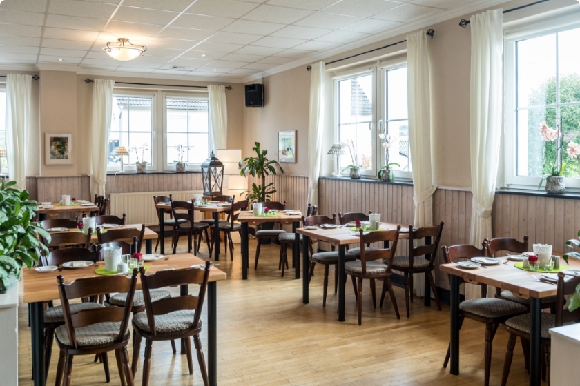 Einblick in das Restaurant des Hotels. Diese ist mit hellem Holzboden ausgestattet und besitzt dunkle Holzstühle sowie Holztische. Im Hintergrund sieht man die Fensterfront mit hellen zusammengebundenenn Gardinen als Dekoelement.