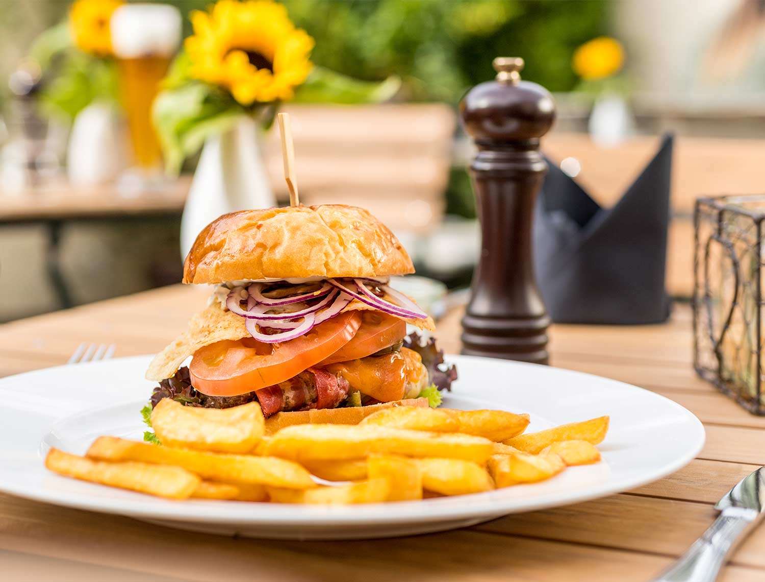 Teller mit einem Burgergericht mit Pommes Frites. Im Burger, der aus Spiegelei, Tomaten, Zwiebeln und  Rinderpatty besteht steckt ein Holzspiess. Im Hintergrund ist eine Dunkle Pfeffermühle sowie Vasen mit Sonnen Blumen zu sehen.