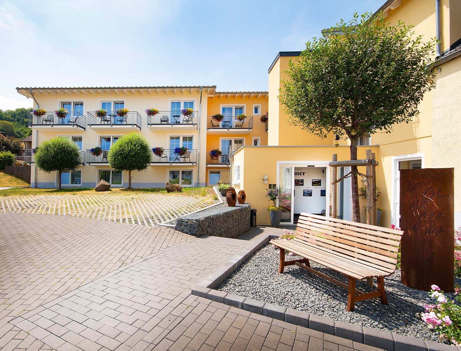 Blick auf den Parkplatz, den Eingang und den Zimmerkomplex. Das Haus ist hellgelb gestrichen.
