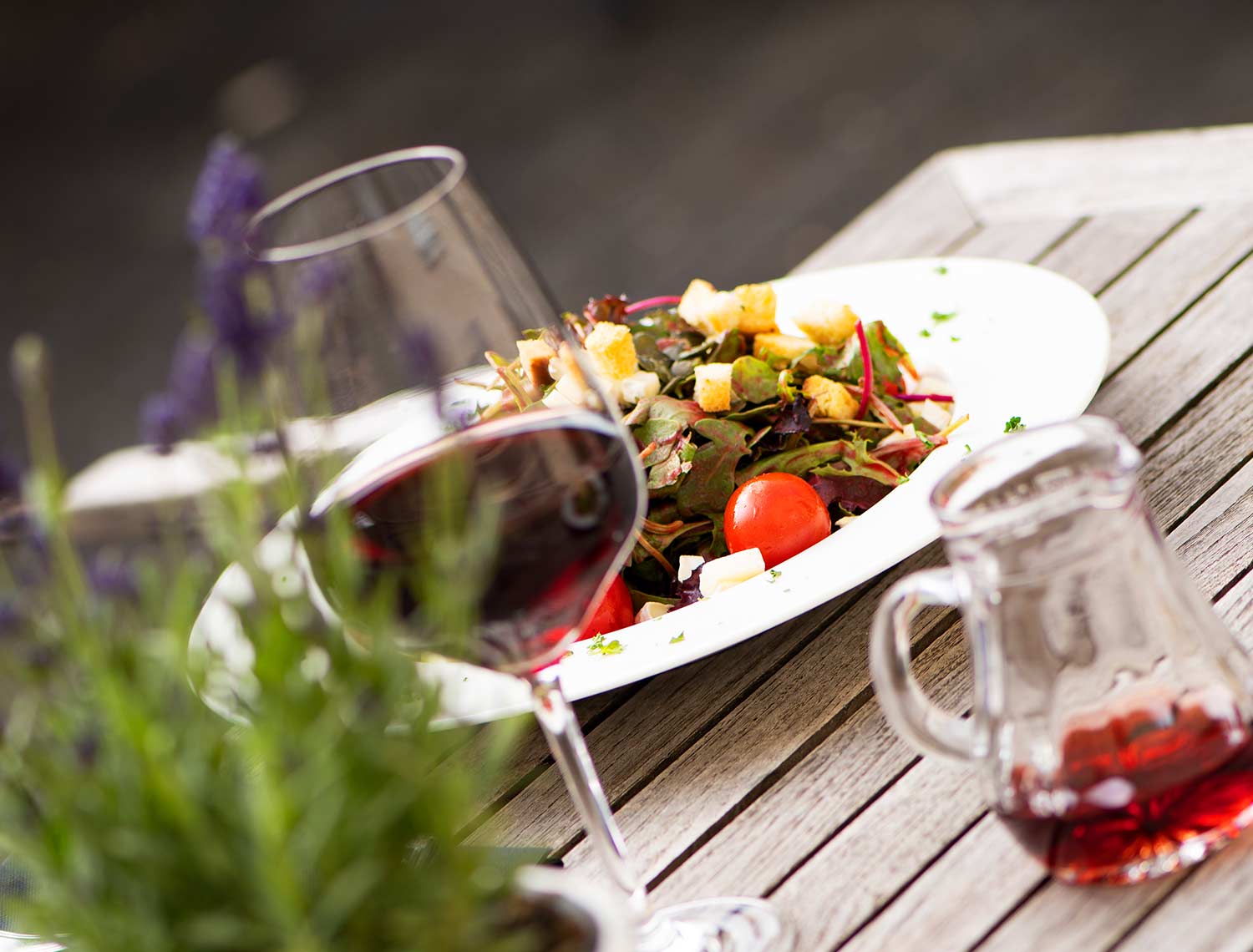 Blick auf einen Bunt gefüllten Teller. Es sind Salate, Croutons und Tomaten zu erkennen. Im Vordergrund sieht man ein Glas Rotwein sowie eine Karaffe mit Rotweinresten.