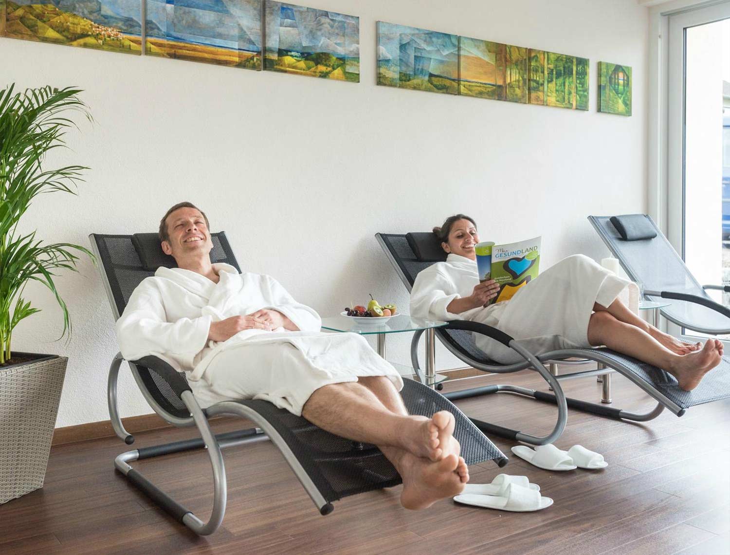 Zwei Personen liegen in den Wellness liegen. Der Herr auf der linken Seite entspannt und die Dame liest in einem Magazin. auf dem Glastisch zwischen den Liegen ist ein Früchteteller.