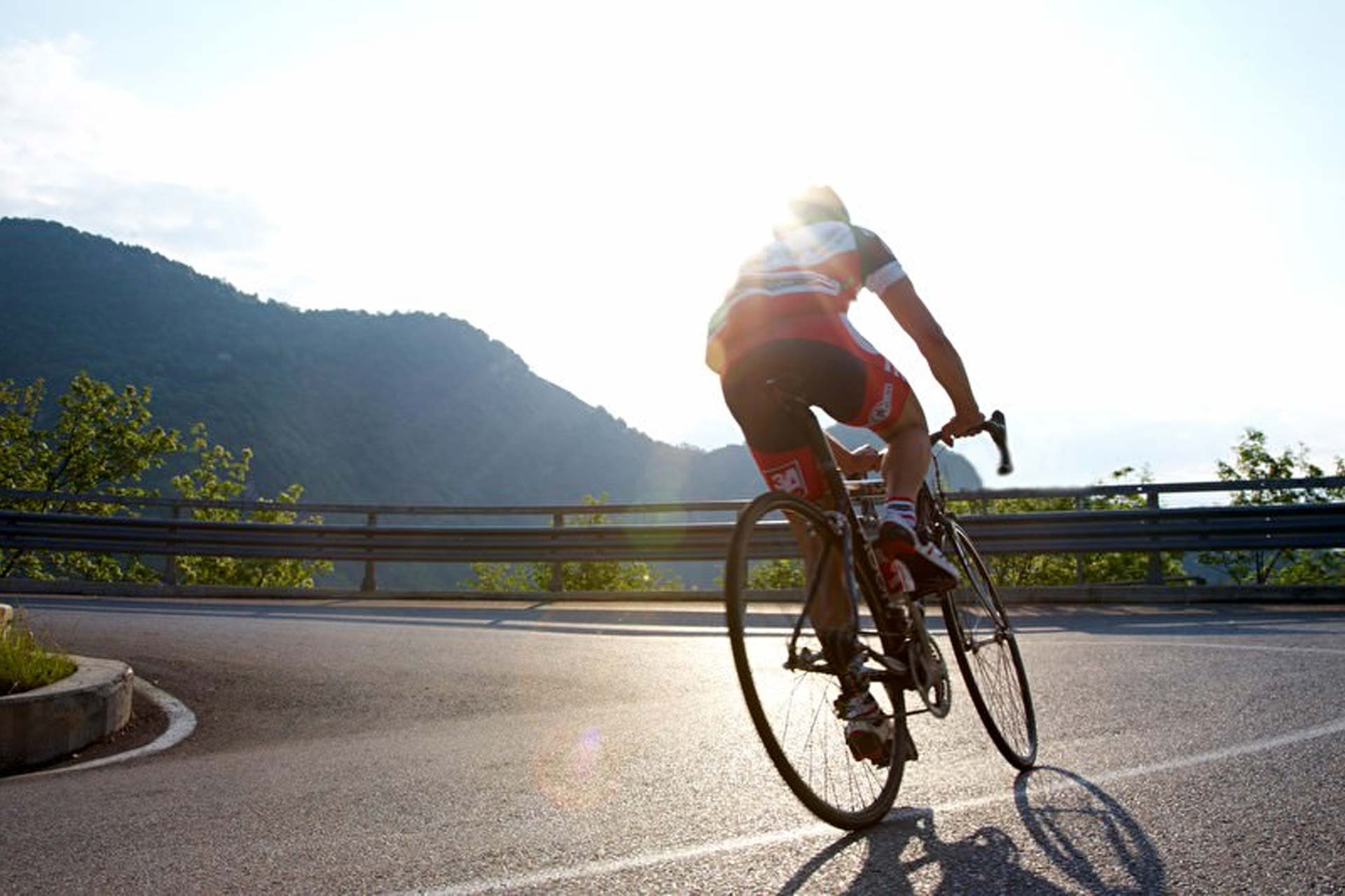 Das Bild zeigt einen Rennradfahrer einen Berg rauf fahren. Im Hintergrund sieht man ein paar Berge.