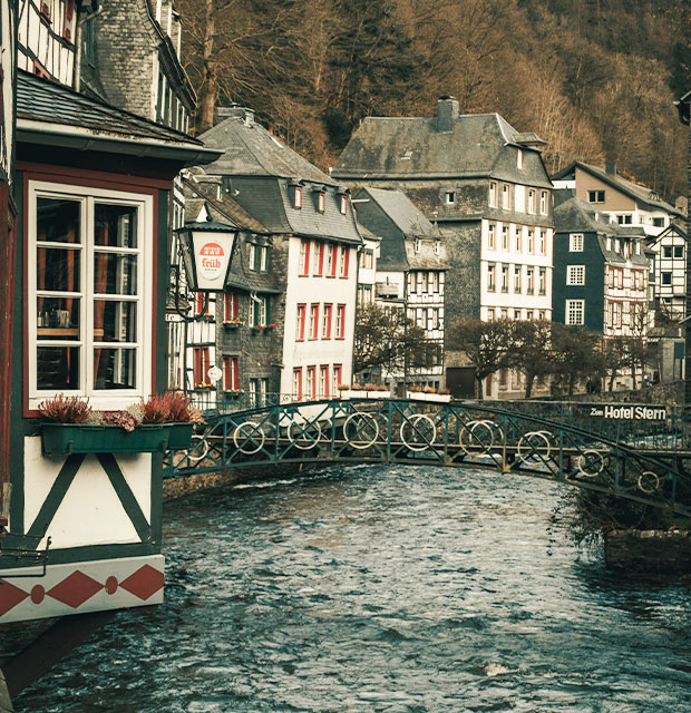 Abbildung der Altstadt Monschau durch die der Fluss Rur fließt. Im Zentrum des Bildes sieht man eine Brücke aus Eisen.