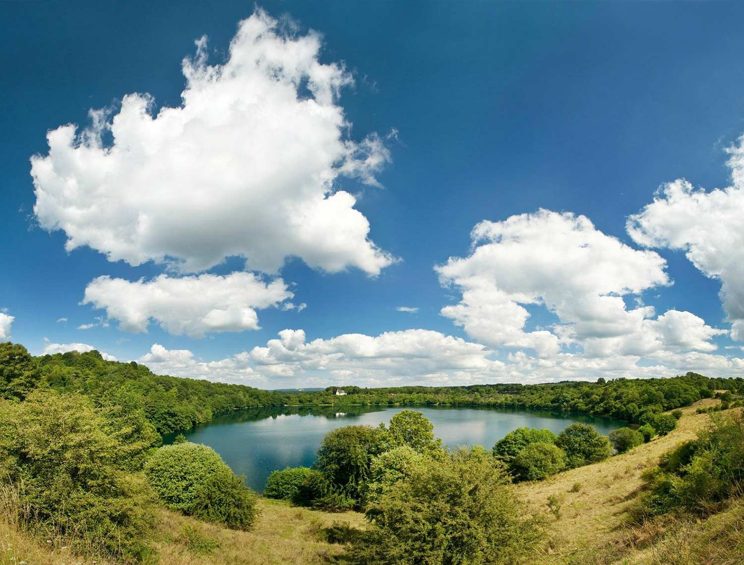 Das Bild ist eine Weitwinkel Aufnahme von einem see mit viel Grünen Bäumen rundherum. Es sind Außerdem sehr viele dicke Wolken am Himmel.