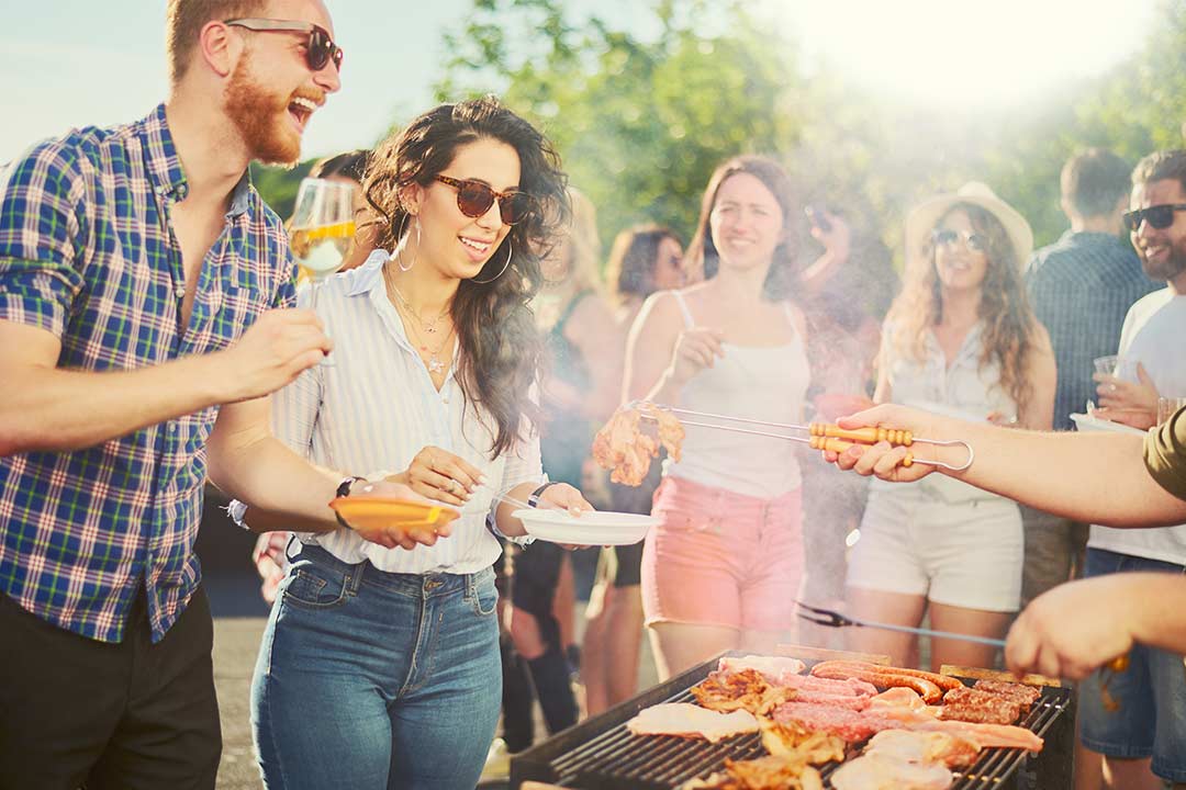 Auf dem Bild ist eine Gruppe von Personen die um einen Tisch stehen. Im Vordergrund sind zwei Personen denen gerade Fleisch hingehalten wird.