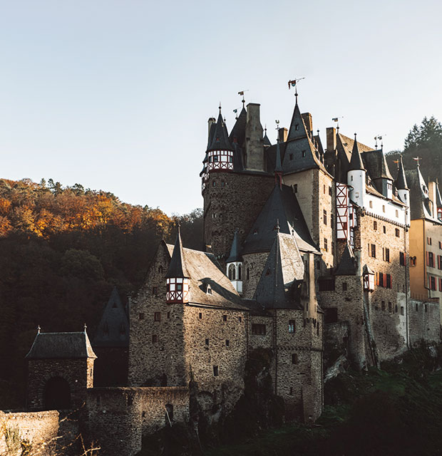 Darstellung der Burg Eltz, in Wierschem, von dessen Eingang aus. Das Bild zeigt die schmal geschnittene Burg mit Ihren vielen Türmen und Erkern sowie der steinernen Brücke die dorthin führt.