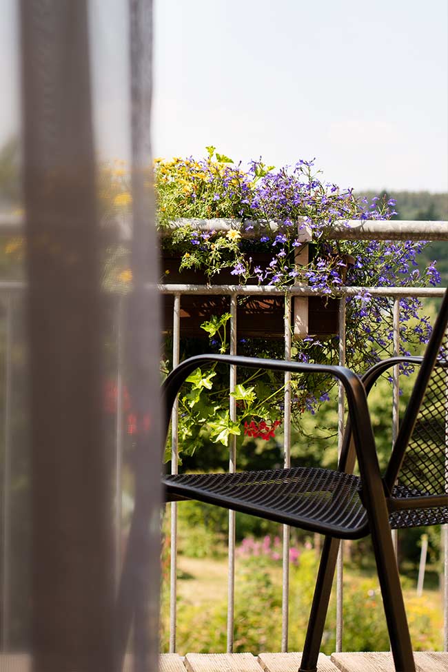 Blick aus dem Zimmer auf den Balkonstuhl und dem bepflanzten Geländer des Balkons. 