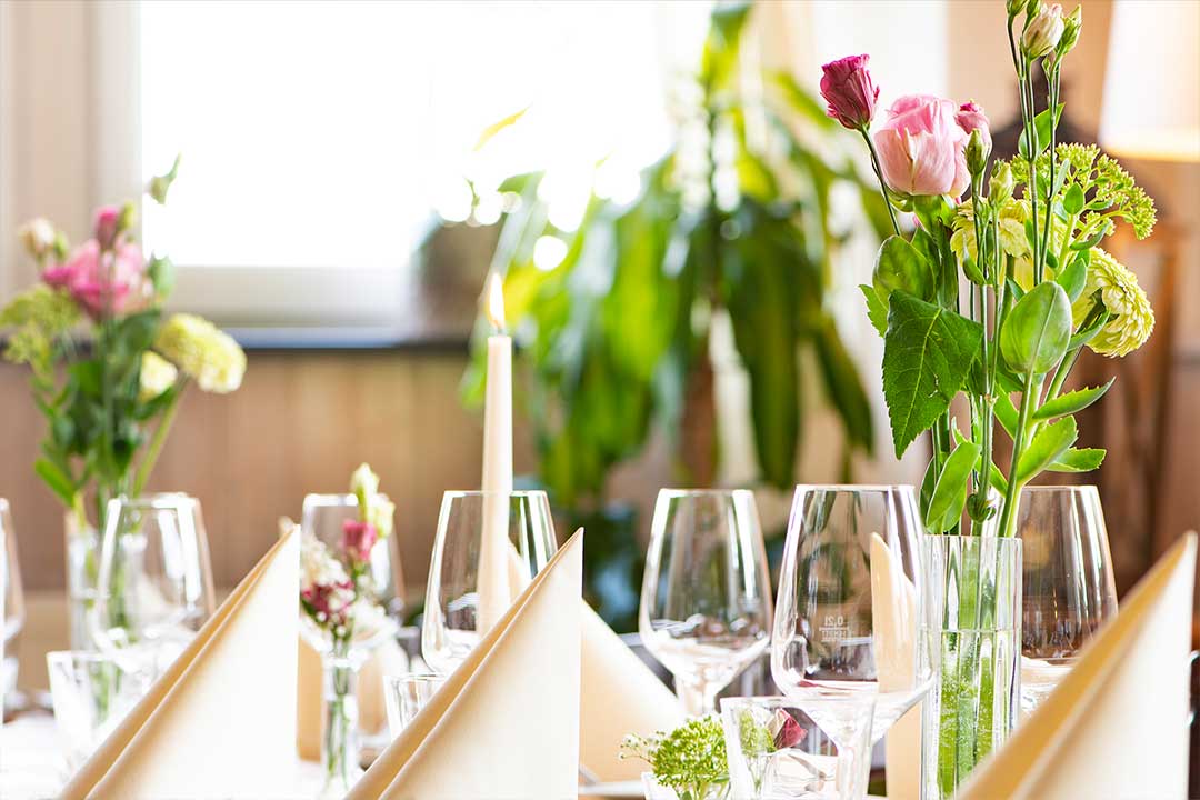 Blichk auf einen gedeckten Tisch . Auf diesem ist eine Vase mit Rosen. zudem sieht man Weißweingläser und eine brennende Kerze