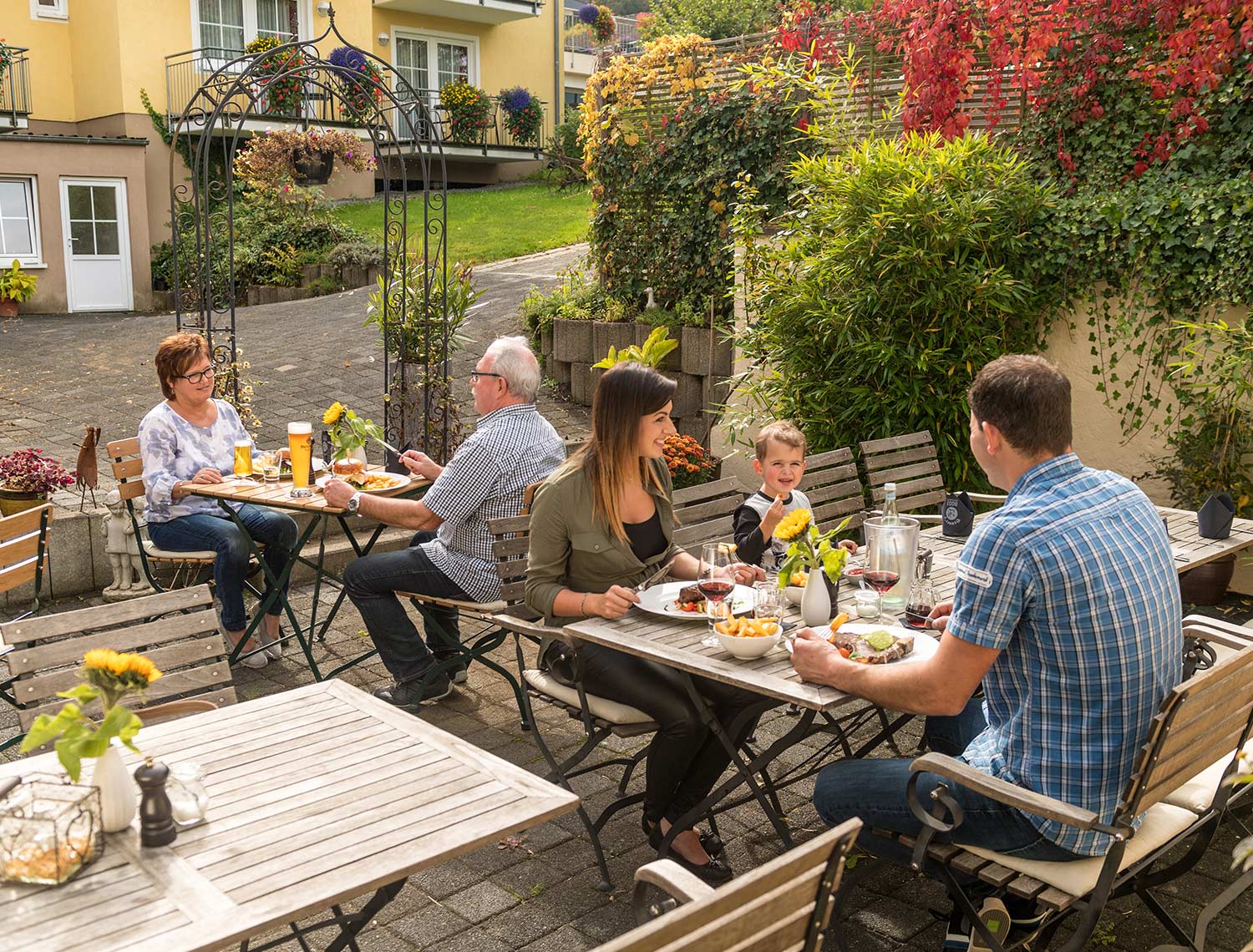 im Vordergrund von dem Bild ist eine Familie (eine Frau ein Mann und ein Kind) zu sehen, welche an einem Gedeckten Tisch sitzen. Im Hintergrund ist ein Altes Ehepaar zu sehen, welche ebenfalls gerade essen. Die ganze scene findet draußen statt und die zwei Familien sitzen an Holz Tischen