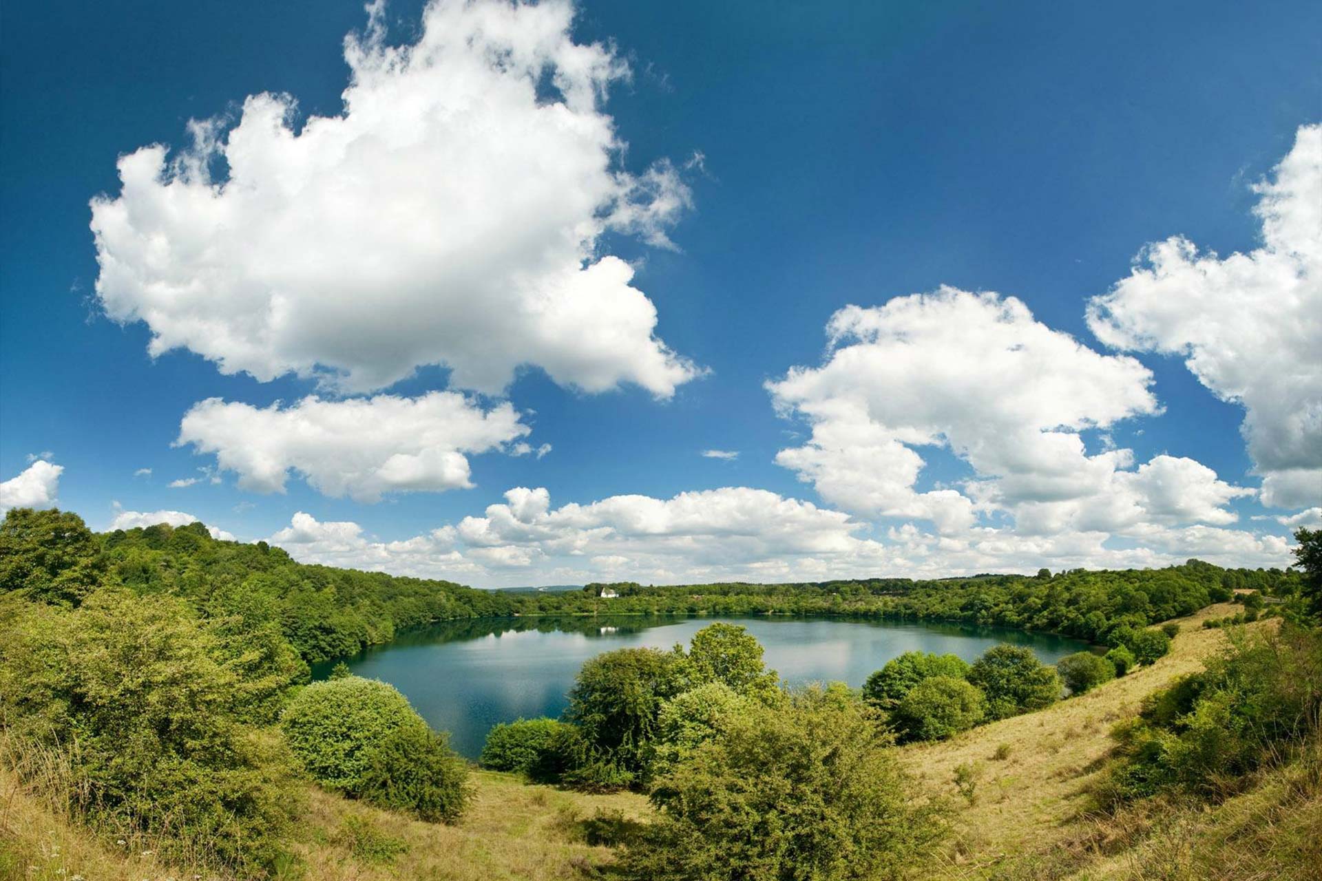 Das Bild ist eine Weitwinkel Aufnahme von einem see mit viel Grünen Bäumen rundherum. Es sind Außerdem sehr viele dicke Wolken am Himmel.