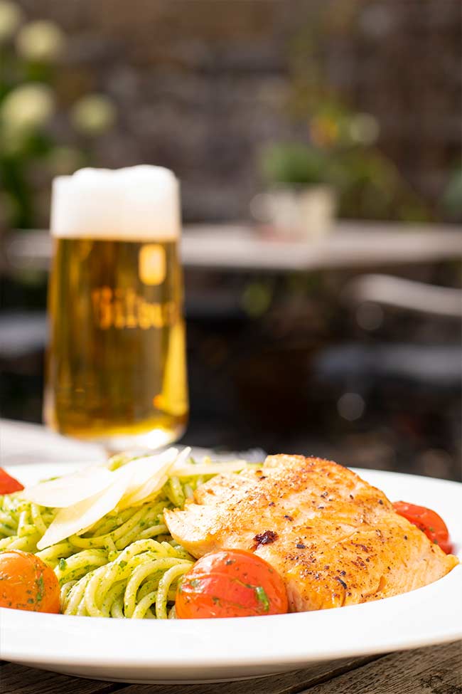 Bild eines Pasta und Lachgerichtes. Die Pasta ist mit grünem Pesto verfeinert. Der Lachs ist an einem Stück. Zudem sind Tomaten zu sehen. Im Hintergrund steht ein Glas Bitburger Bier.